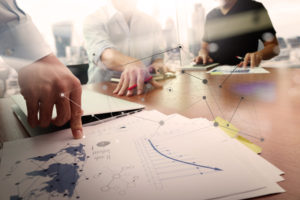 business documents on office table with smart phone and laptop computer and graph business with social network diagram and two colleagues discussing data in the background